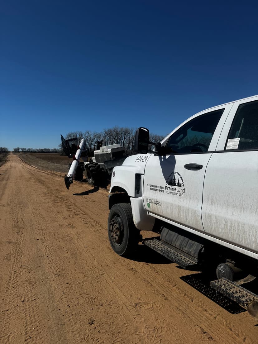 White truck on a dirt road near a farming implement under a clear blue sky.